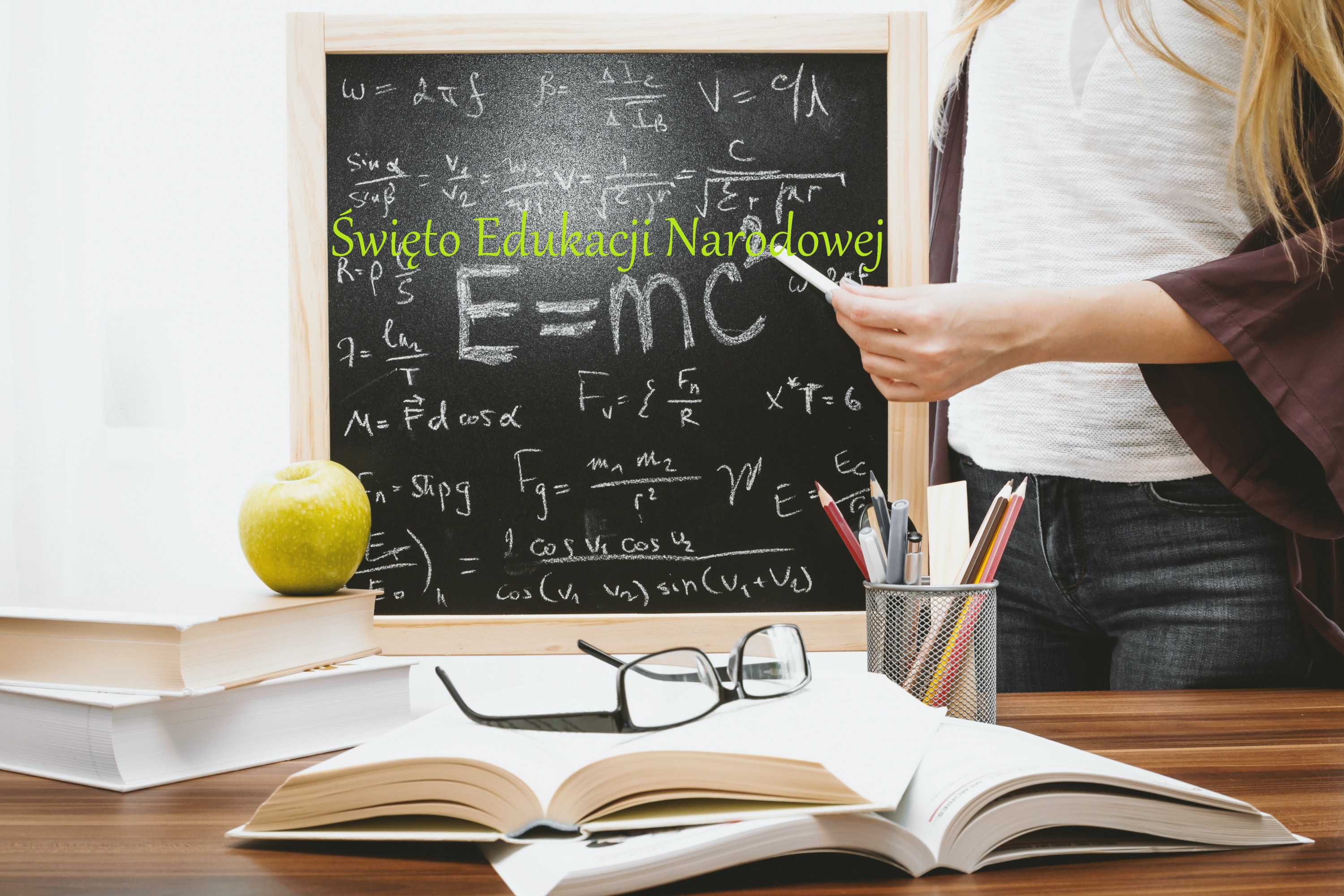 Tablica, na tablicy napis Święto Edukacji Narodowej, widać postać nauczycielki trzymającej kredę, na biurku książki przybory do pisania, jabłko